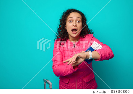 Surprised stunned woman with open mouth, surprised expression, checking time, fear of being late for the flight, standing near her suitcase and holding her passport with air ticket and boarding pass 85380806