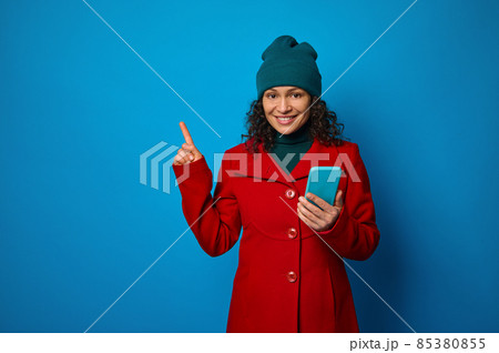 Conceptual portrait of a cheerful pretty woman wearing bright red coat and warm green woolen hat holding mobile phone and pointing with her finger on a copy space for ads, isolated on blue background 85380855