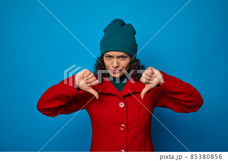Disappointed upset sad frustrated pretty woman in bright red coat and green hat looking at camera and showing thumbs up, posing against blue background with copy ad space 85380856