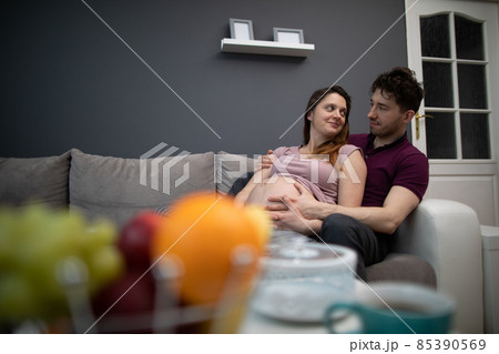 A married couple looks at each other with great joy while sitting on the sofa in the living room. The woman is pregnant. A married couple looks at each other with great joy while sitting on the sofa in the living room. The woman is pregnant. 85390569