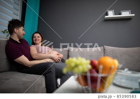 A married couple looks at each other with great joy while sitting on the sofa in the living room. The woman is pregnant. A married couple looks at each other with great joy while sitting on the sofa in the living room. The woman is pregnant. 85390583