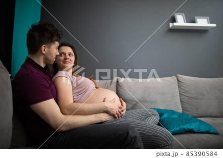 A married couple looks at each other with great joy while sitting on the sofa in the living room. The woman is pregnant. A married couple looks at each other with great joy while sitting on the sofa in the living room. The woman is pregnant. 85390584