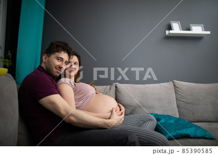 A married couple huddled sitting on a sofa looking straight into the lens. Pregnant woman. 85390585