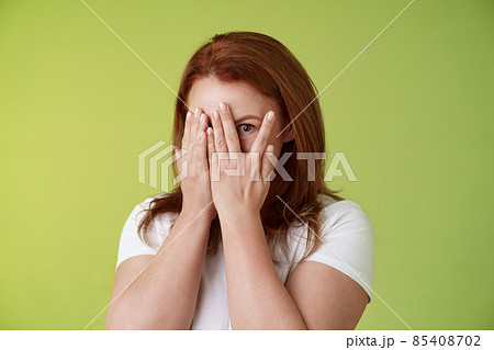 Not peeking. Playful charismatic middle-aged ginger redhead woman close face palms look through fingers one eye stare camera intrigued waiting surprise gift stand green background 85408702