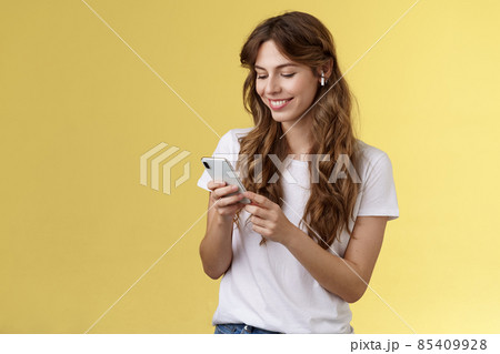 Happy caring tender feminine girlfriend curly long hair hold smartphone picking song listen way home smiling broadly look mobile phone scroll music platform wear wireless earbud yellow background Happy caring tender feminine girlfriend curly long hair hold smartphone picking song listen way home smiling broadly look mobile phone scroll music platform wear wireless earbud yellow background 85409928