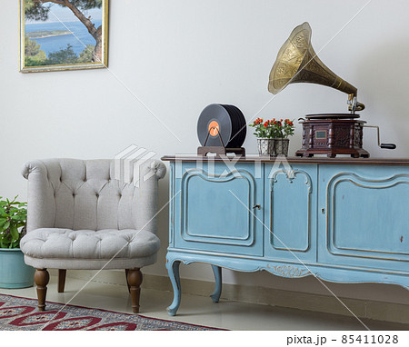 Retro off white armchair, vintage wooden light blue sideboard, old phonograph (gramophone) and vinyl records on background of beige wall, tiled porcelain floor, and red carpet Retro off white armchair, vintage wooden light blue sideboard, old phonograph (gramophone) and vinyl records on background of beige wall, tiled porcelain floor, and red carpet 85411028