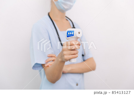 Temperature measurement gun in doctor hands. Close-up shot of doctor wearing protective surgical mask ready to use infrared isometric thermometer gun to check body temperature for virus symptoms. Temperature measurement gun in doctor hands. Close-up shot of doctor wearing protective surgical mask ready to use infrared isometric thermometer gun to check body temperature for virus symptoms. 85413127