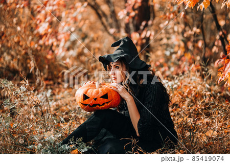 Sitting girl in witch costume holding pumpkin in her hands in front of the forest 85419074