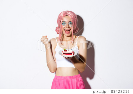 Portrait of cheerful smiling girl celebrating her birthday, wearing pink wig, holding b-day cake and shouting of joy, standing over white background 85428215