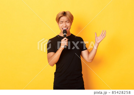 Handsome asian male singer, korean guy singing song at karaoke in microphone with passion, standing over yellow background Handsome asian male singer, korean guy singing song at karaoke in microphone with passion, standing over yellow background 85428258