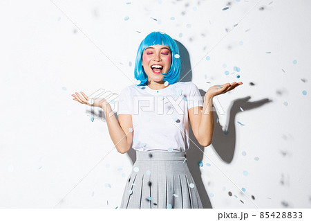 Portrait of happy asian girl in blue wig celebrating halloween, throwing confetti in the air, standing over white background 85428833