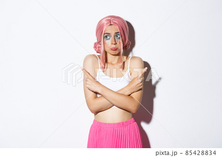 Portrait of clueless silly young girl in pink wig and halloween costume, shrugging and pouting indecisive, pointing fingers sideways, standing over white background 85428834
