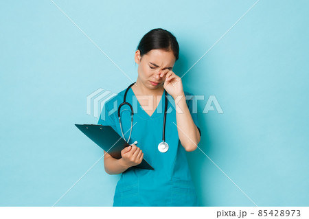Covid-19, social distancing and coronavirus pandemic concept. Overworked exhausted asian female doctor, nurse in scrubs rubbing tired eyes during shift in clinic, holding clipboard Covid-19, social distancing and coronavirus pandemic concept. Overworked exhausted asian female doctor, nurse in scrubs rubbing tired eyes during shift in clinic, holding clipboard 85428973
