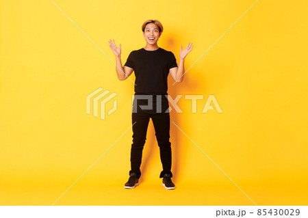 Full-length of surpirsed and excited, smiling blond asian guy raising hands up amazed, yellow background 85430029