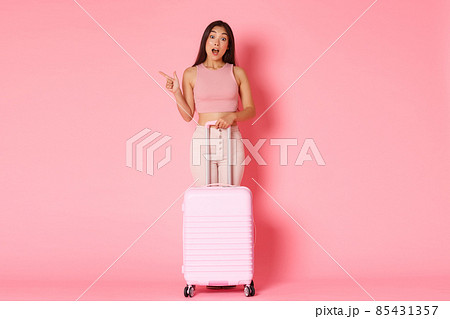 Travelling, holidays and vacation concept. Full-length of curious and wondered asian beautiful girl tourist pointing finger left, asking question, standing with suitcase over pink background Travelling, holidays and vacation concept. Full-length of curious and wondered asian beautiful girl tourist pointing finger left, asking question, standing with suitcase over pink background 85431357