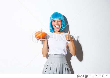 Happy cute asian girl celebrating halloween, wearing party costume and wig, holding small pumpkin, showing thumbs-up in approval 85432423