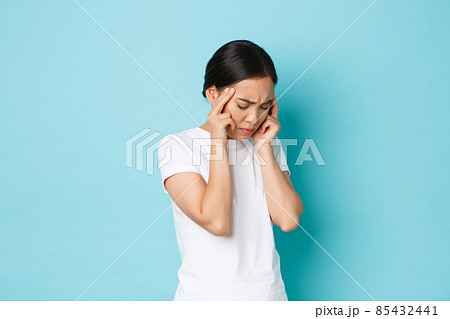 Side view of bothered tired asian girl grimacing, touching head and complaining on headache, having pain in head, migraine of feeling dizzy, standing blue background sick Side view of bothered tired asian girl grimacing, touching head and complaining on headache, having pain in head, migraine of feeling dizzy, standing blue background sick 85432441