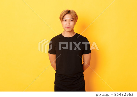 Portrait of handsome smiling asian blond guy looking sassy, holding hands behind back, standing yellow background Portrait of handsome smiling asian blond guy looking sassy, holding hands behind back, standing yellow background 85432962