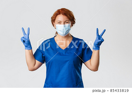 Medical workers, covid-19 pandemic, coronavirus concept. Optimistic smiling redhead female doctor, physician in medical mask and gloves showing peace gesture, staying positive during quarantine Medical workers, covid-19 pandemic, coronavirus concept. Optimistic smiling redhead female doctor, physician in medical mask and gloves showing peace gesture, staying positive during quarantine 85433819