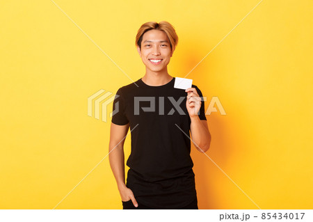 Cheerful handsome asian guy smiling pleased and showing credit card, standing yellow background 85434017