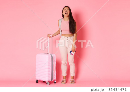 Travelling, holidays and vacation concept. Full-length of crying, sad and distressed asian girl tourist, feeling depressed about missed flight, standing suitcase and plane tickets, pink background 85434471