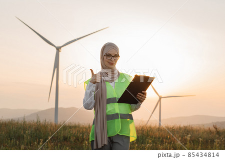 Woman in hijab standing on windmill farm with clipboard Woman in hijab standing on windmill farm with clipboard 85434814