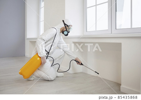 Pest control exterminator getting rid of insects inside infested house. Man in protective mask and suit spraying insecticide from yellow bottle on floor and walls for safe living environment at home 85435868