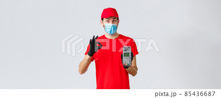 Contactless delivery, payment and online shopping during covid-19, self-quarantine. Excited courier in red uniform, face mask and gloves recommend pay order with POS terminal and credit card Contactless delivery, payment and online shopping during covid-19, self-quarantine. Excited courier in red uniform, face mask and gloves recommend pay order with POS terminal and credit card 85436687