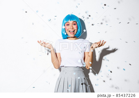 Portrait of happy asian girl in blue wig celebrating halloween, throwing confetti in the air, standing over white background 85437226