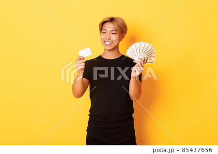 Portrait of satisfied smiling, handsome asian guy looking pleased at credit card while also holding money, standing yellow background Portrait of satisfied smiling, handsome asian guy looking pleased at credit card while also holding money, standing yellow background 85437656