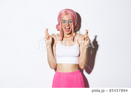 Image of optimistic party girl in pink wig, with halloween makeup, close eyes and screaming excited while cross fingers to make a wish, standing over white background 85437894
