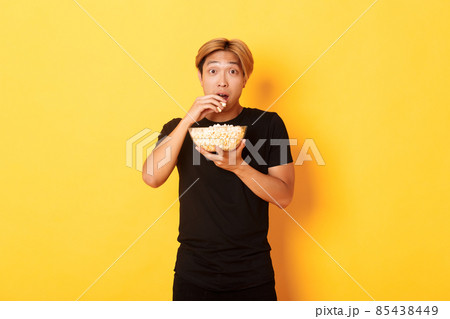 Excited and amazed asian guy watching interesting movie and eating popcorn from bowl, standing yellow background 85438449