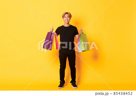 Full-length of attractive korean guy in black clothes, showing thumbs-up, holding shopping bags, yellow background 85438452