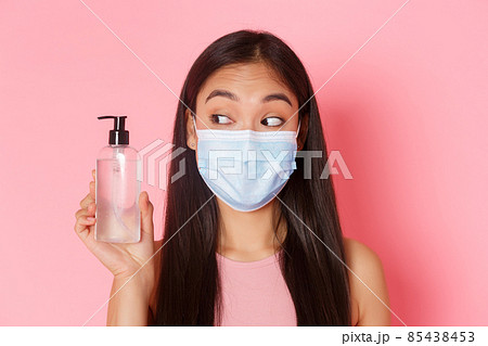 Covid-19 pandemic, coronavirus and social distancing concept. Close-up of cute and silly, pretty asian girl in medical mask looking at hand sanitizer, advice using antiseptics, pink background Covid-19 pandemic, coronavirus and social distancing concept. Close-up of cute and silly, pretty asian girl in medical mask looking at hand sanitizer, advice using antiseptics, pink background 85438453