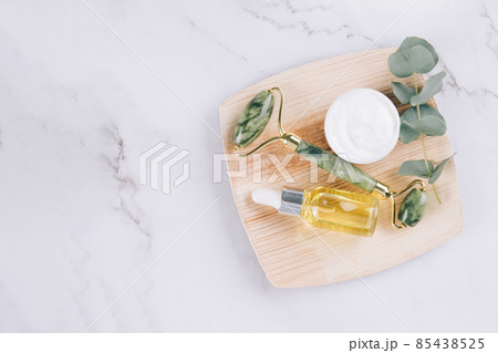 Top view on jade facial roller and gua sha tool with beauty serum, face oil and face moisturizer on white marble table background. Facial massage kit for lifting massage therapy, home spa. Mock up 85438525