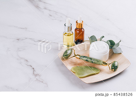Jade facial roller and gua sha tool with beauty serum, face oil and face moisturizer on white marble table background. Facial massage kit for lifting massage therapy, home spa. Mock up 85438526