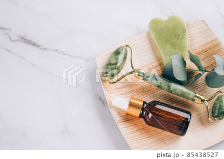 Jade gua sha facial roller with beauty serum oil on white marble table background with copy space. Facial massage kit for lifting massage therapy made from natural stones 85438527