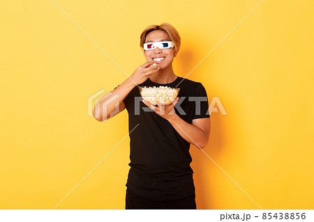 Joyful handsome asian guy with blond hair, watching movie or tv series in 3d glasses, eating popcorn and smiling amused, standing yellow background 85438856