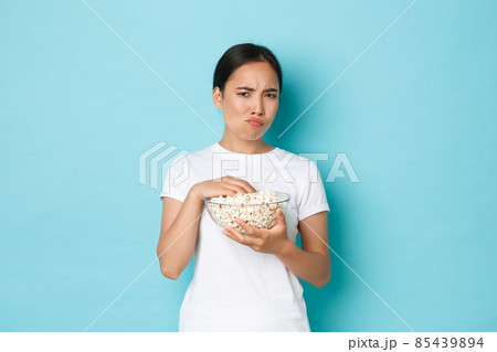 Lifestyle, leisure and emotions concept. Skeptical and displeased asian cute girl watching movie and eating popcorn, grimacing from cringe content, looking suspicious, standing light blue background 85439894