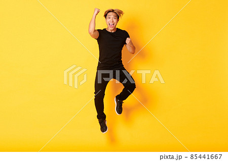 Full-length of happy attractive asian guy in black clothes jumping and celebrating victory, achieve goal, standing yellow background Full-length of happy attractive asian guy in black clothes jumping and celebrating victory, achieve goal, standing yellow background 85441667