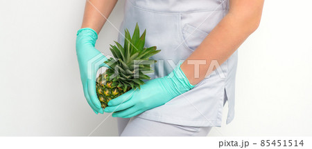 Young beautician wearing blue gloves in uniform with pineapple covers an intimate area on a white background, bikini zone depilation concept. Young beautician wearing blue gloves in uniform with pineapple covers an intimate area on a white background, bikini zone depilation concept. 85451514