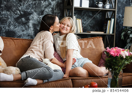 Mother and daughter relationship concept. A woman and a teenage girl are sitting in front of the TV on the couch and have fun talking. The family spends time at home. Little dog lying on laps Mother and daughter relationship concept. A woman and a teenage girl are sitting in front of the TV on the couch and have fun talking. The family spends time at home. Little dog lying on laps 85454406