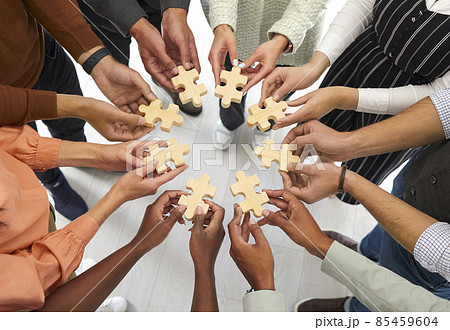 Business and cooperation. Multiracial people stand in circle and connect wooden pieces of puzzles they hold in their hands. Cropped image of hands holding puzzle pieces symbolizing unity and teamwork. 85459604