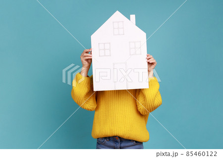 Portrait of unknown little kid hiding behind paper house, affordable housing program, advertisement, wearing yellow casual style sweater. Indoor studio shot isolated on blue background. Portrait of unknown little kid hiding behind paper house, affordable housing program, advertisement, wearing yellow casual style sweater. Indoor studio shot isolated on blue background. 85460122