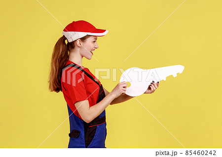 Side view portrait of young happy satisfied worker woman holding big paper key in hands, looking ahead, wearing overalls and red cap. Indoor studio shot isolated on yellow background. Side view portrait of young happy satisfied worker woman holding big paper key in hands, looking ahead, wearing overalls and red cap. Indoor studio shot isolated on yellow background. 85460242