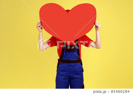Portrait of optimistic worker woman hiding her face behind big red heart, expressing romantic feelings, wearing overalls and red cap. Indoor studio shot isolated on yellow background. 85460284