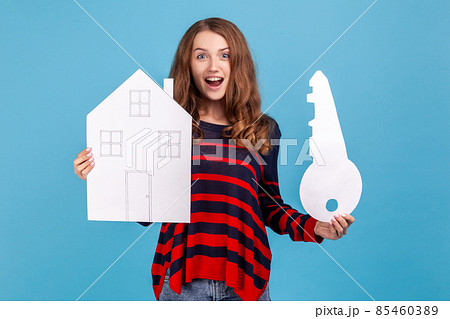 Surprised astonished woman wearing striped casual style sweater, holding paper house and big key, real estate purchase, rental services, mortgage. Indoor studio shot isolated on blue background. 85460389