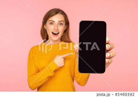 Portrait of amazed good looking girl in sweater pointing at cellphone and looking with surprise at camera, shocked about device or mobile application. indoor studio shot pink background 85460392