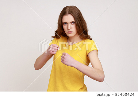 Determined teenager girl with brown hair with fighting spirit in casual yellow T-shirt punching with fists, boxing, looking confidently at camera. Indoor studio shot isolated on gray background. Determined teenager girl with brown hair with fighting spirit in casual yellow T-shirt punching with fists, boxing, looking confidently at camera. Indoor studio shot isolated on gray background. 85460424