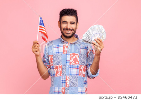 Happy smiling man with beard wearing blue casual style shirt looking at camera, holding banknotes and USA flag in hands, expressing positive emotions. Indoor studio shot isolated on pink background. Happy smiling man with beard wearing blue casual style shirt looking at camera, holding banknotes and USA flag in hands, expressing positive emotions. Indoor studio shot isolated on pink background. 85460473